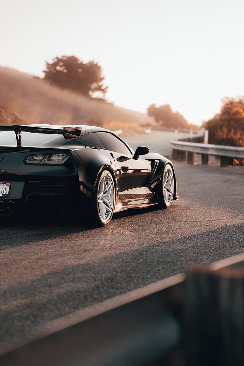 Black Corvette Stingray with silver wheels drives on a winding road during a golden sunset over hazy hills.