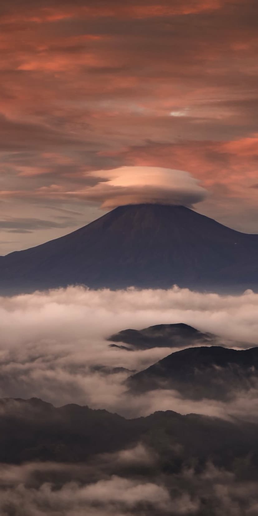 Mount Fuji peak topped with a circular lenticular cloud above pink-tinged morning mist and rolling foothills.