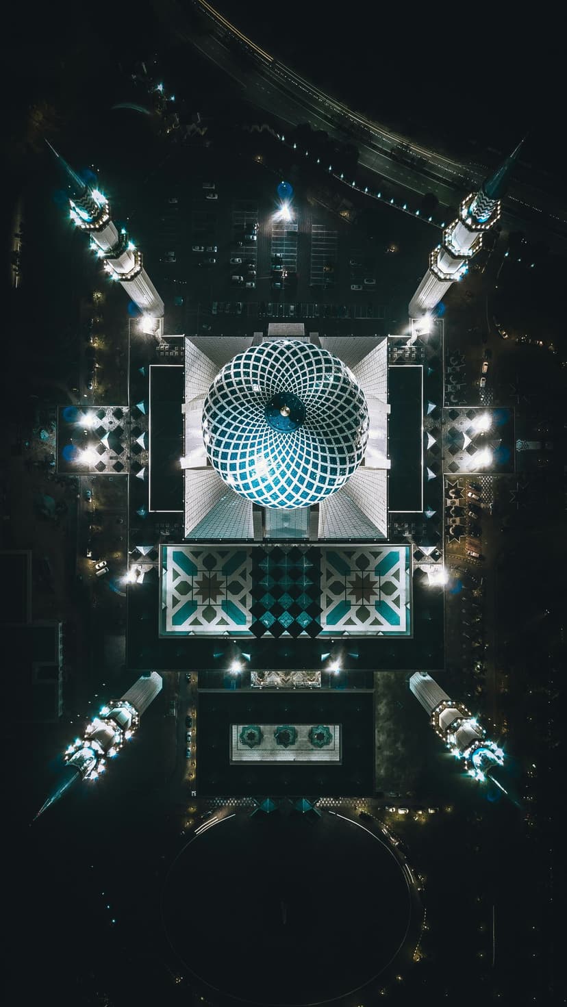 Sultan Salahuddin Abdul Aziz Shah Mosque dome and minarets glow with blue and gold lights at night from above.