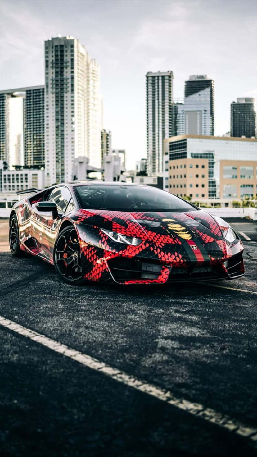 Red and black snake-patterned Lamborghini parked on an urban street with tall skyscrapers in the background.