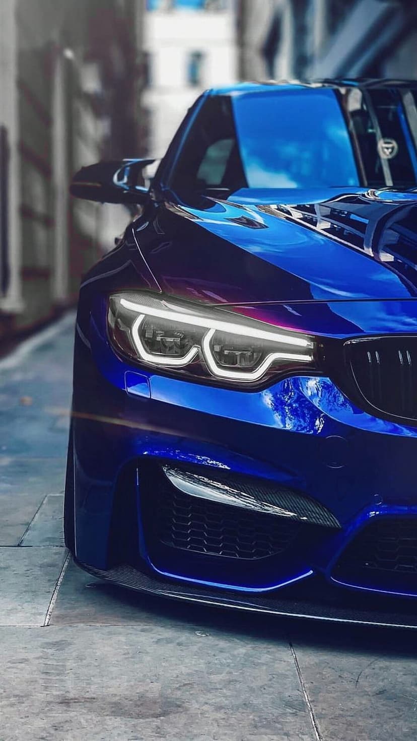 BMW M4 front end featuring blue metallic paint, glowing LED headlights, and a black kidney grille in close-up.