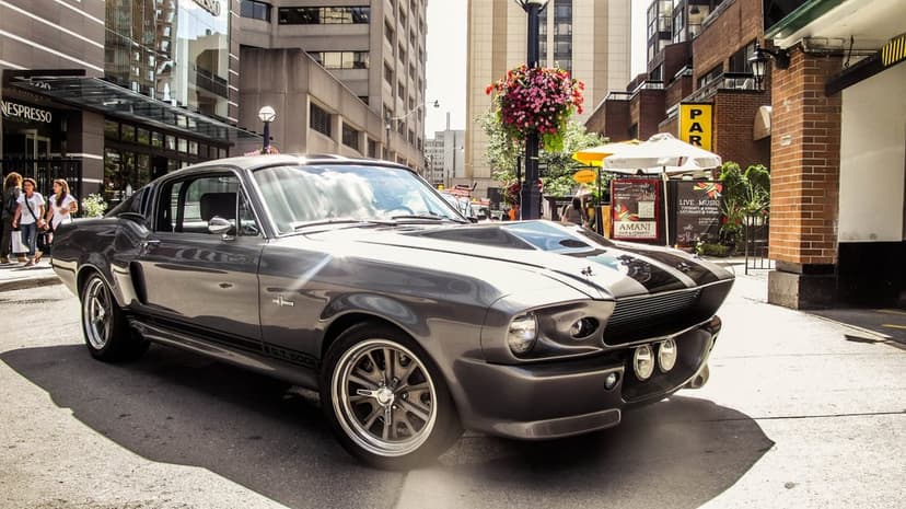 Gray Shelby GT500 Eleanor Mustang with black stripes parked on a sunlit city street with urban buildings.
