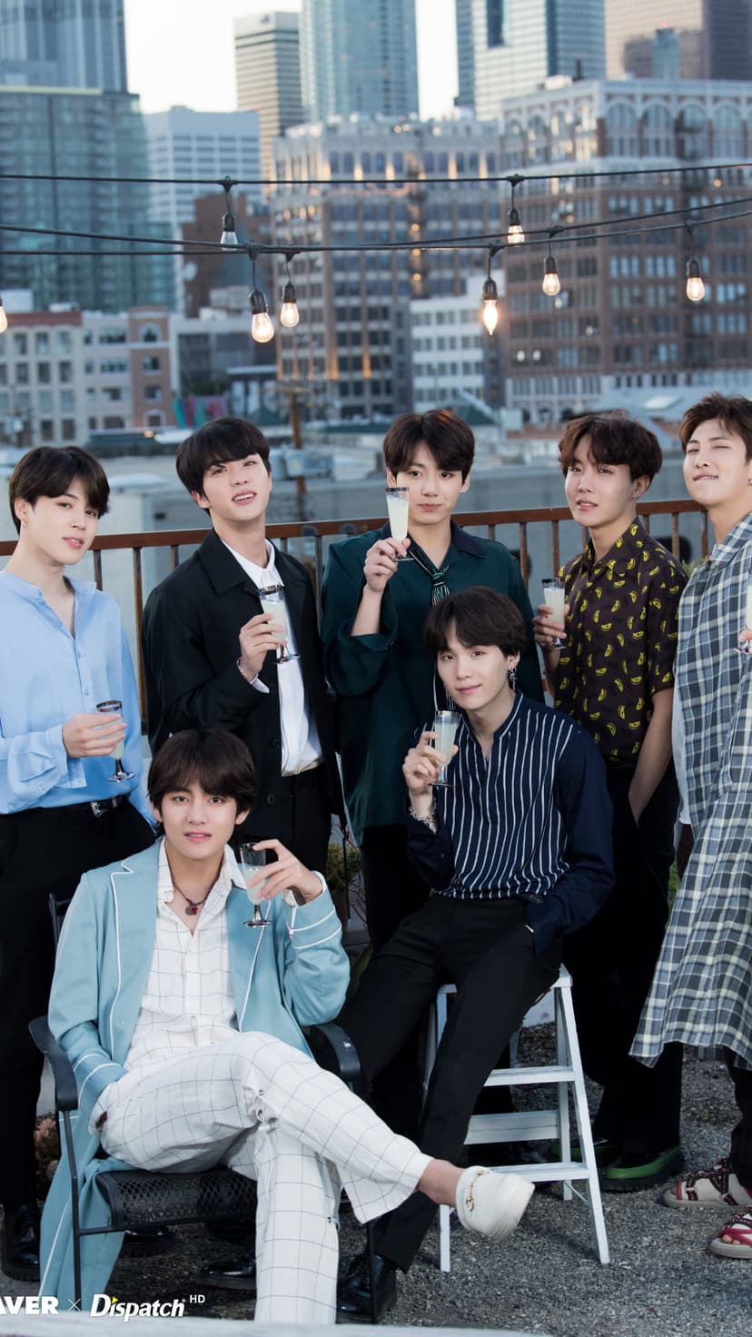 BTS members holding glasses on a rooftop with a blurred city skyline backdrop during a sunset group celebration.