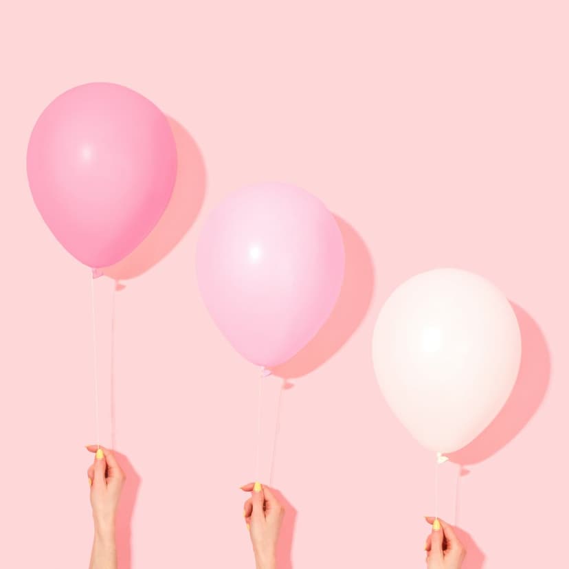 Three pastel balloons in shades of pink and cream held by hands against a matching soft pink studio background.
