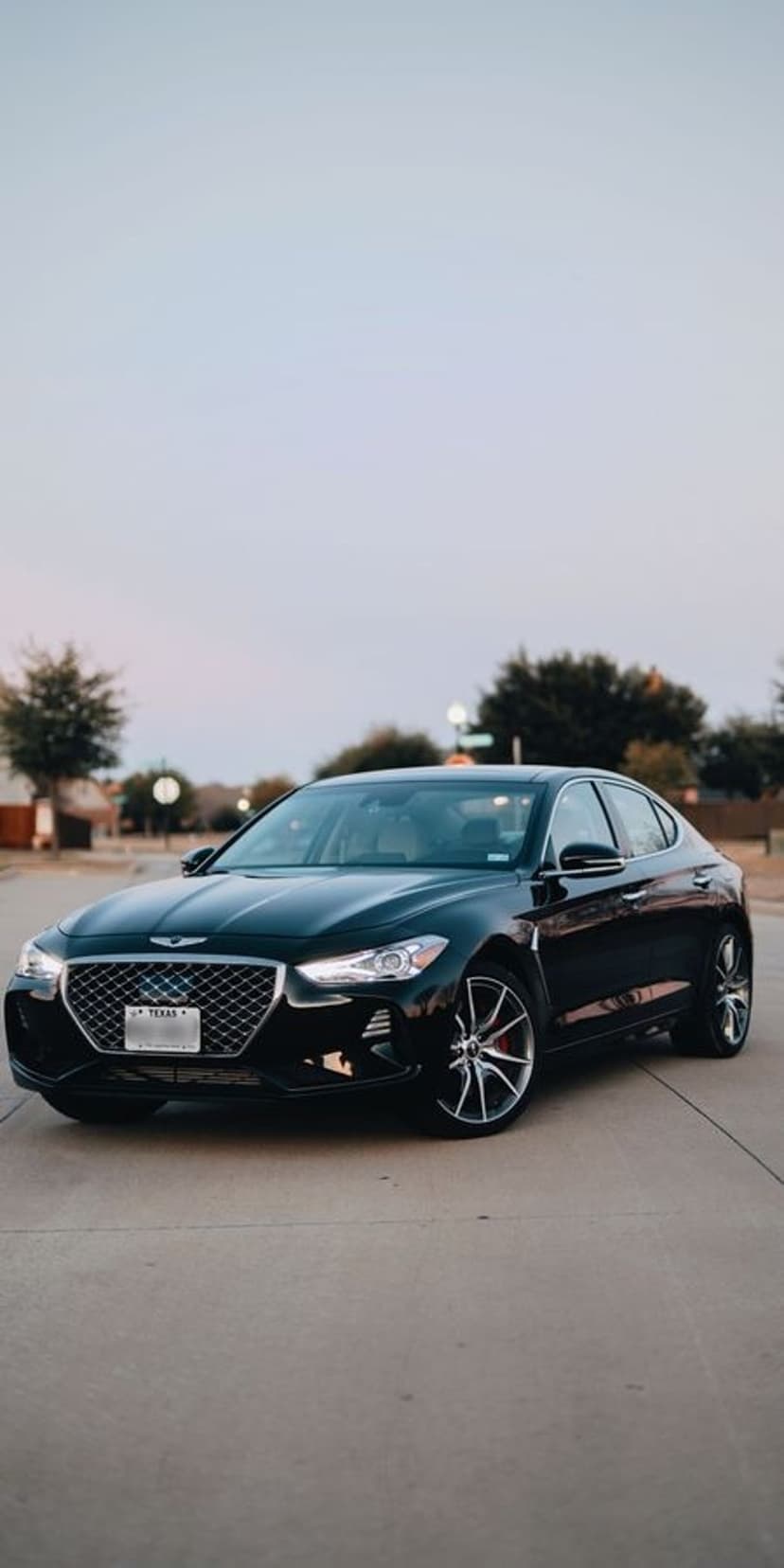 Black Genesis G70 luxury sedan with quad LED headlights parked on a suburban street under a twilight sky.