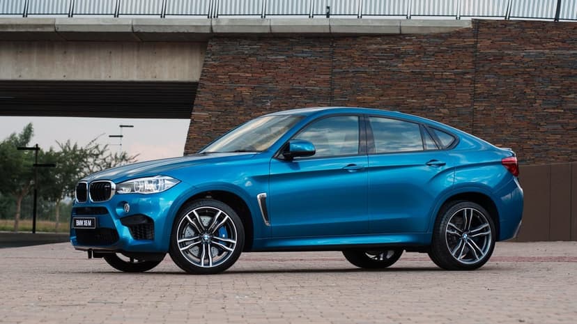 Blue BMW X6 M luxury SUV parked on a paved lot in front of a textured stone wall and concrete bridge.