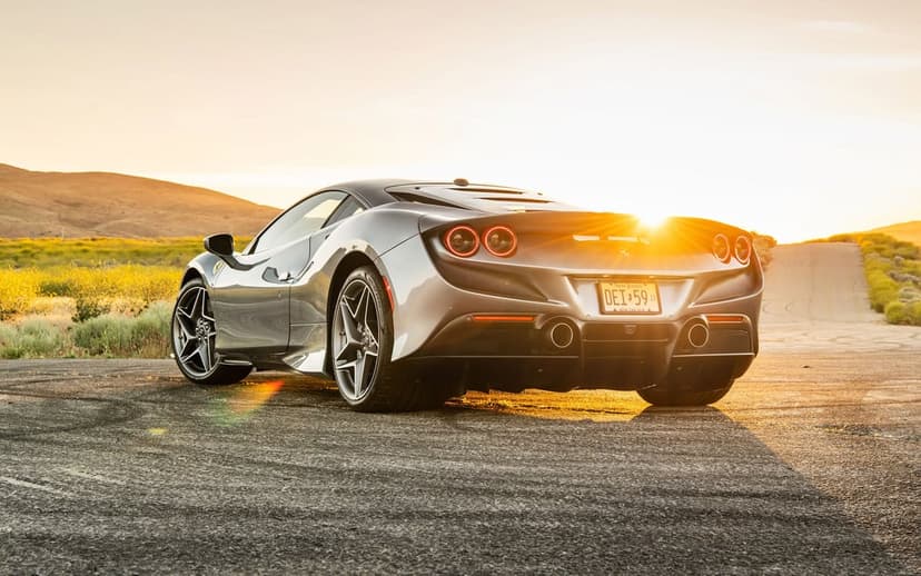 Silver Ferrari F8 Tributo parked on a paved road during a golden sunset with hills and a bright lens flare.
