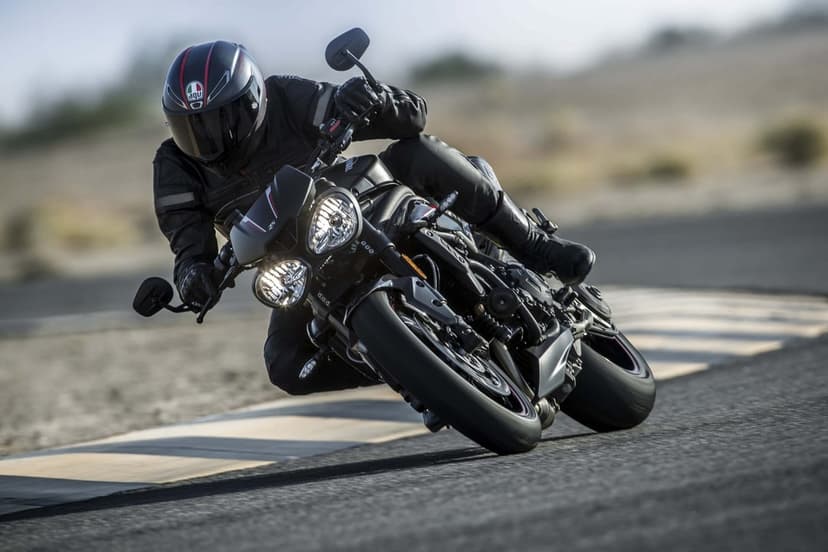Black Triumph motorcycle leaning into a track turn with a rider in matching gear and helmet on grey asphalt.