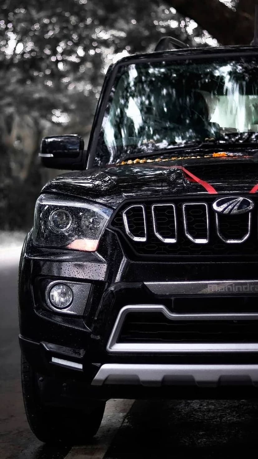 Black Mahindra Scorpio SUV front grille and headlights covered in raindrops against a blurred forest background.