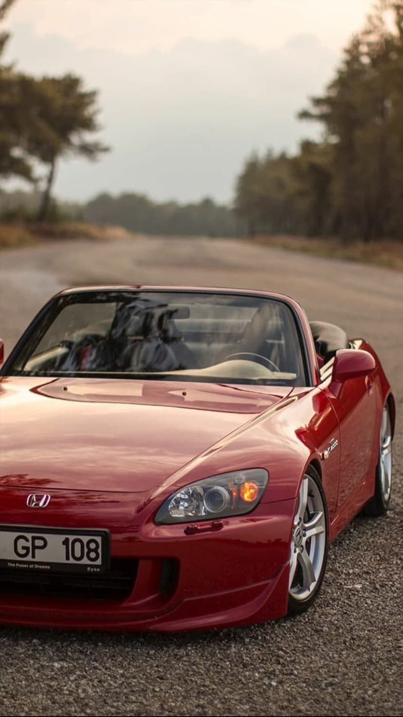 Red Honda S2000 convertible parked on a gray gravel road with a soft-focus forest background and green trees.