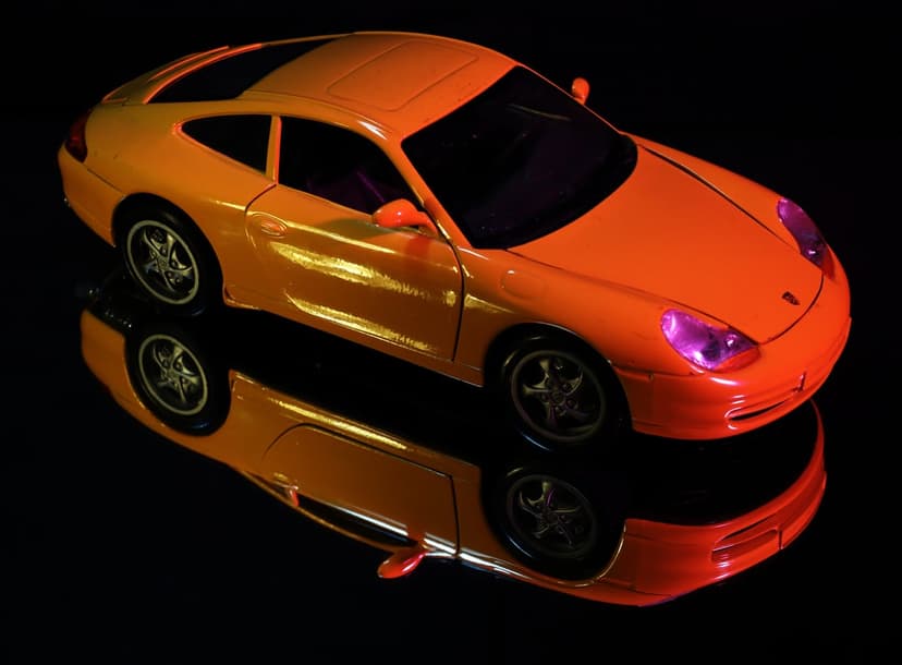 Orange Porsche toy car with a metallic finish reflects on a glossy black surface under bright studio lighting.