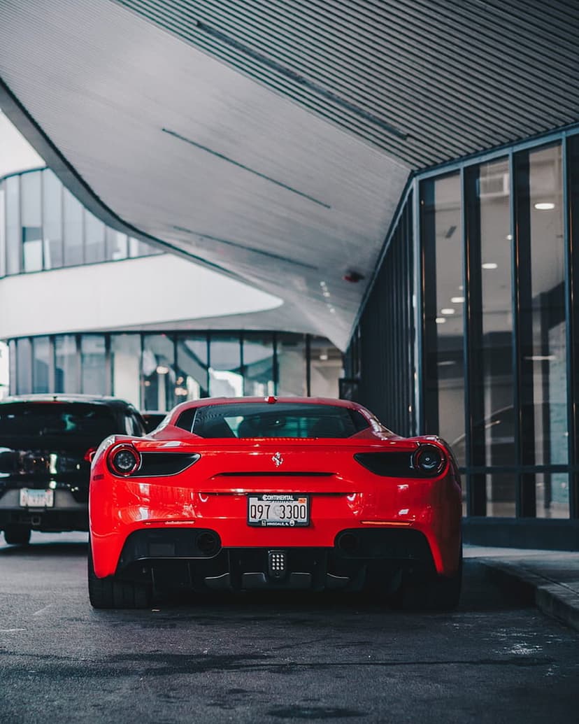Red Ferrari sports car rear view parked beside a modern glass building on an urban street at a low angle.