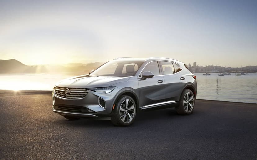 Silver luxury SUV parked on asphalt near a calm bay with city buildings and a glowing sunset in the distance.