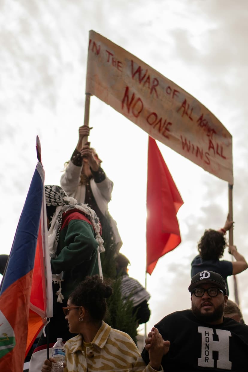 Protesters holding a large banner reading In the war of all against all, no one wins amid a crowd with flags.