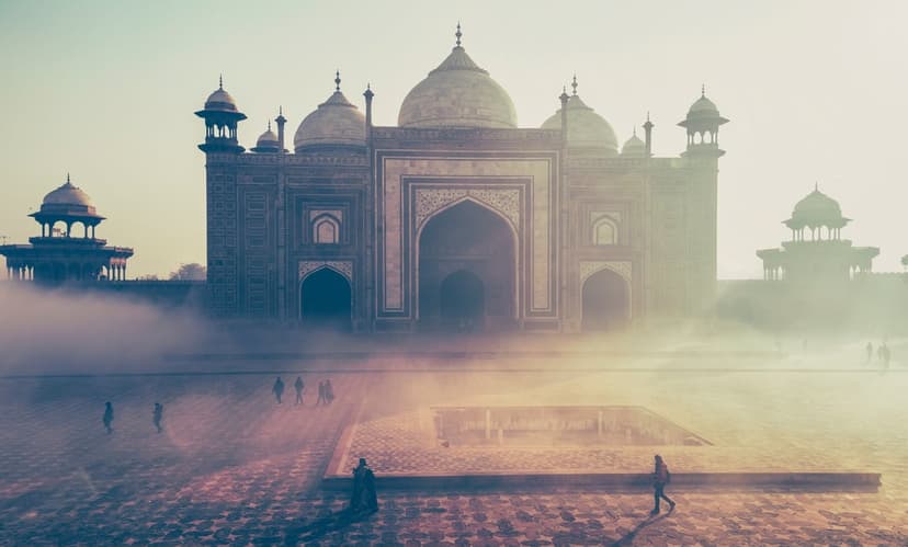 Taj Mahal mausoleum and minarets silhouetted against a hazy sunrise with small figures in the foreground.