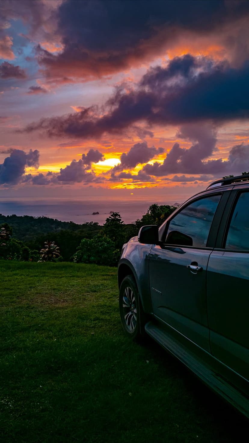 Dark SUV parked on a grassy ridge overlooking a colorful tropical ocean sunset with distant islands.