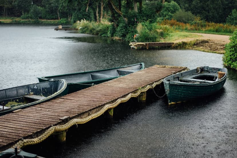 Dark green rowboats tied to a rustic wooden dock on a rippling lake during a gentle rain under an overcast sky.