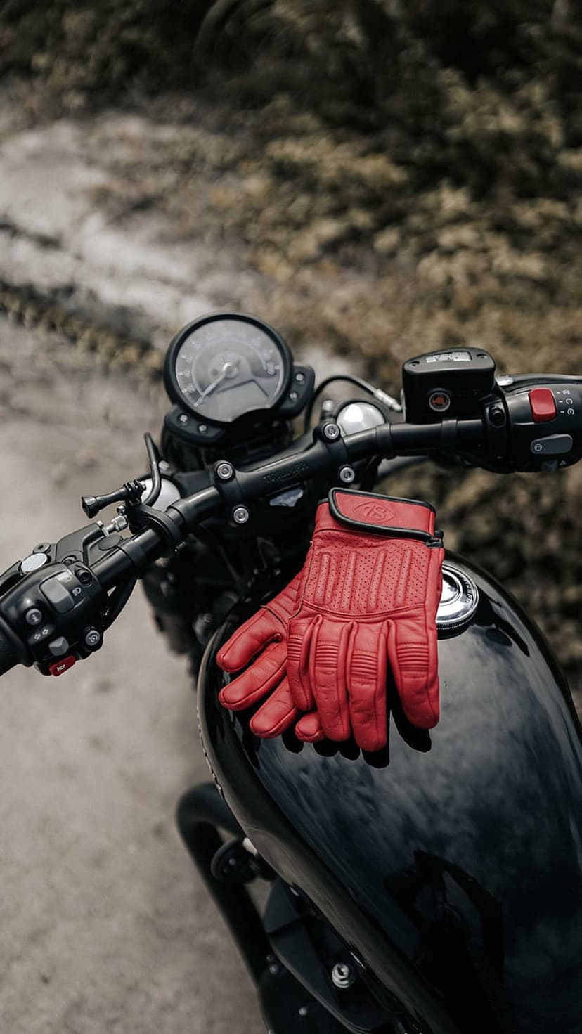 Red leather motorcycle gloves lie on a glossy black tank with a vintage speedometer in the soft-focus background.