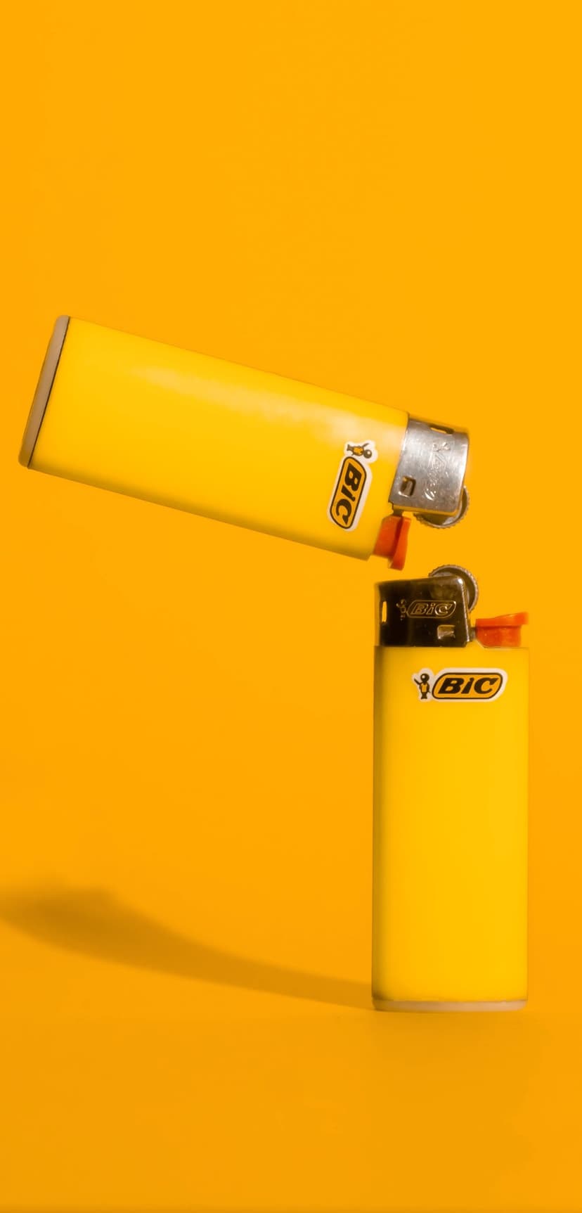 Two yellow butane lighters with chrome caps positioned on a vibrant orange background with soft shadows.