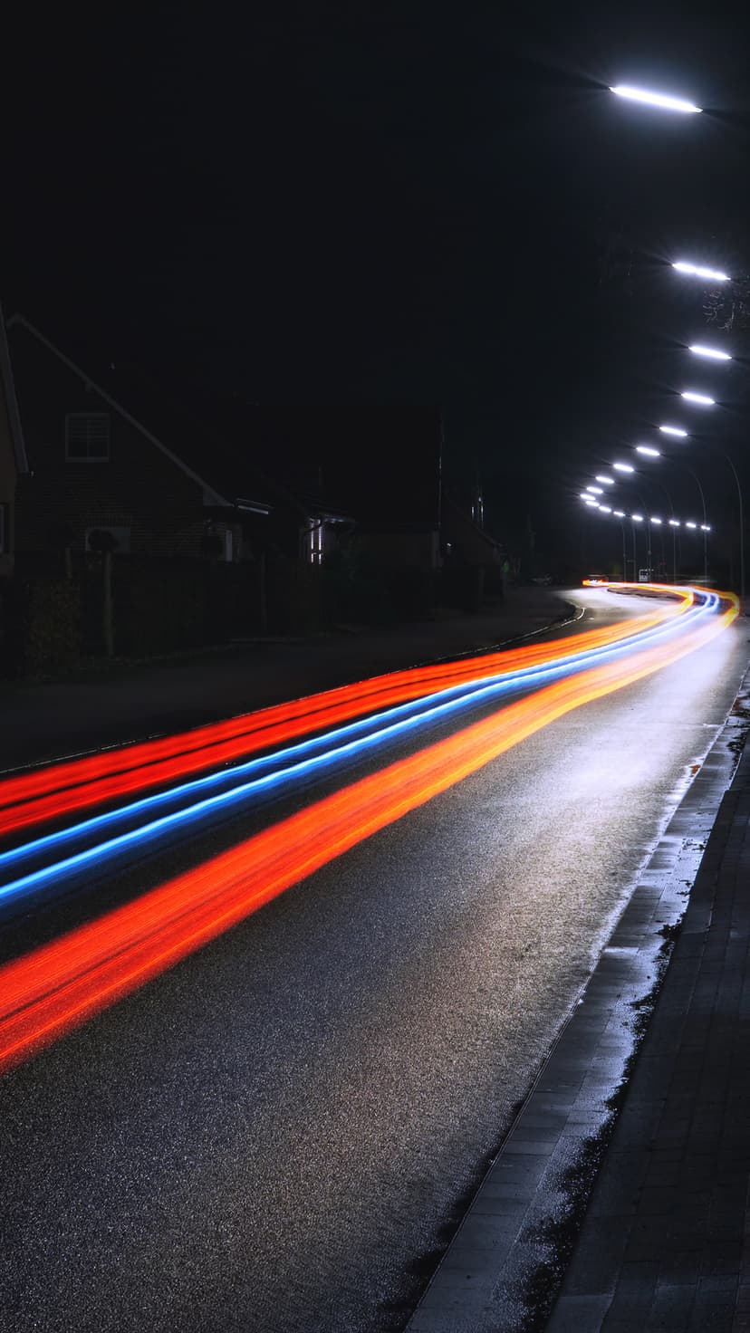 Vibrant red and blue light trails stretch along a dark urban road lined with glowing streetlights at night.