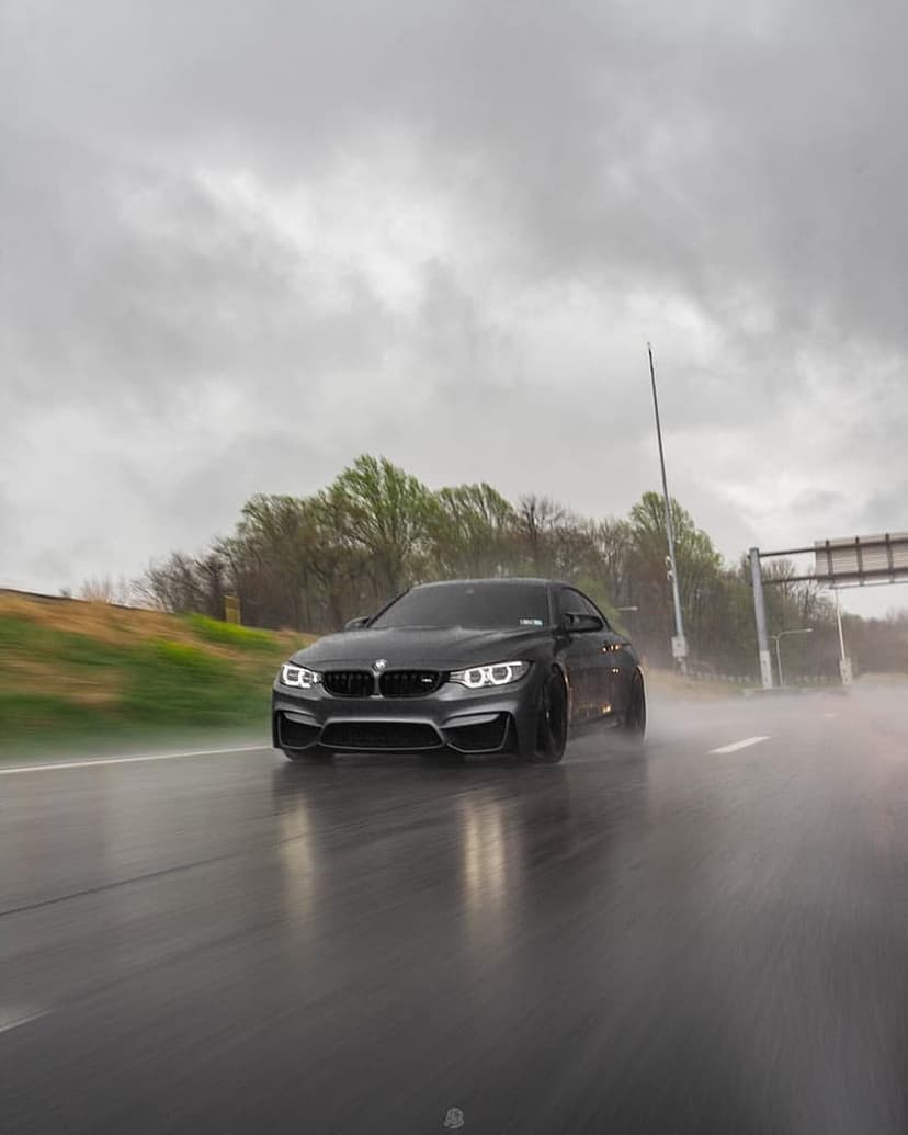 BMW M4 sports car speeds along a wet highway lined with green trees under a dark, dramatic overcast sky.