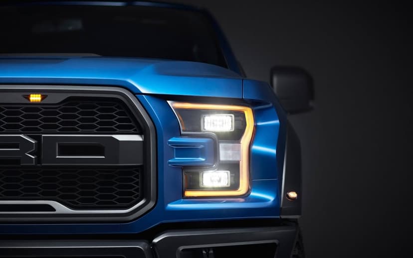 Close-up of a blue Ford Raptor truck's LED headlight and black honeycomb grille under dramatic studio lighting.