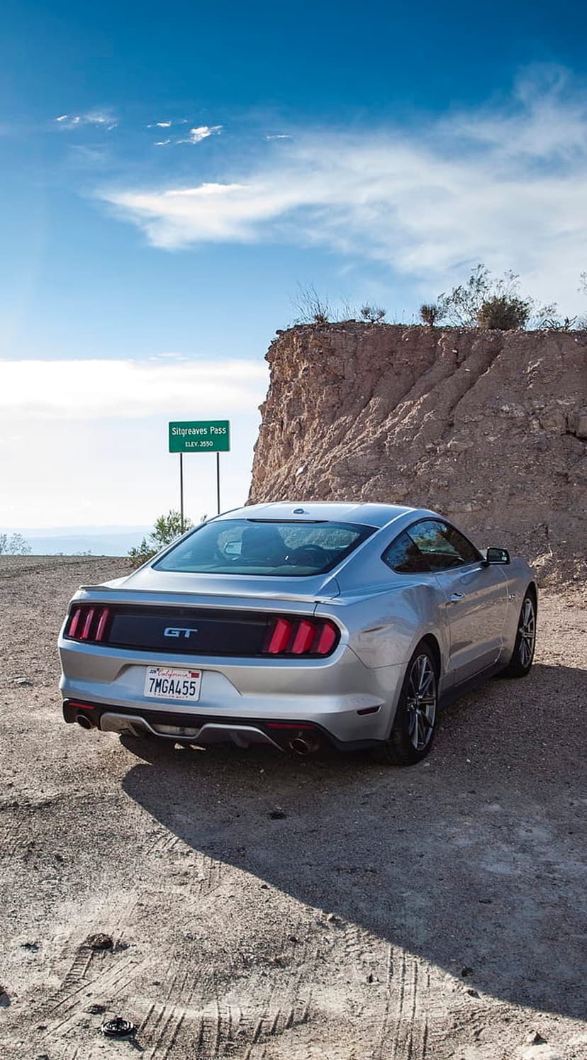 Silver Ford Mustang GT parked on a dusty road beside a rocky cliff and Sitgreaves Pass sign under a blue sky.