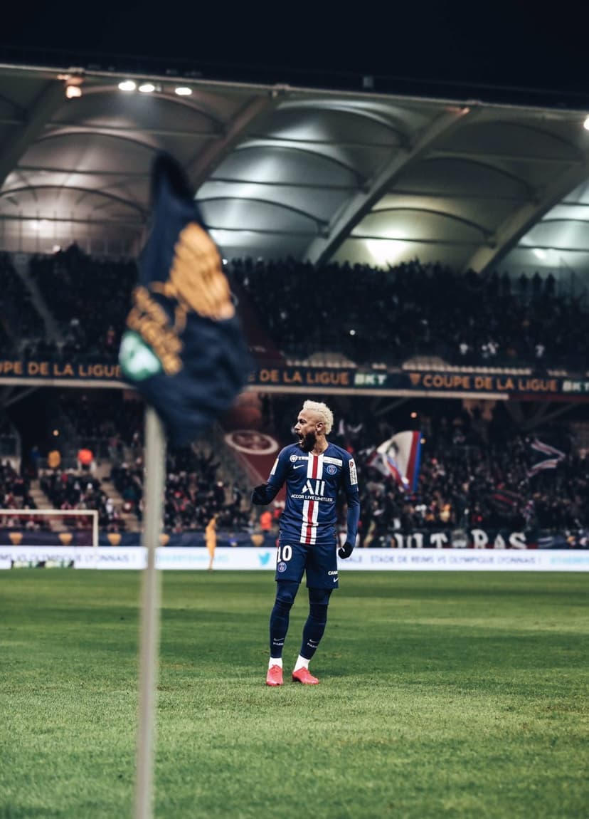 Neymar Jr stands on a grass football pitch at night wearing a navy blue Paris Saint-Germain kit under stadium lights.