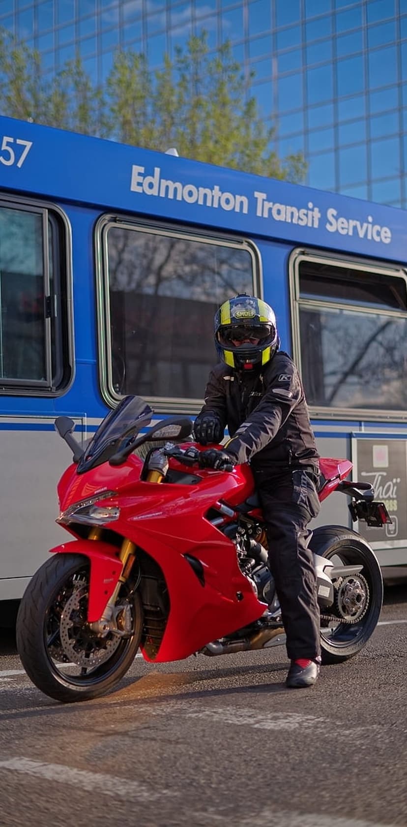 Red Ducati sportbike rider parked near a blue transit bus in front of a reflective glass building in daylight.