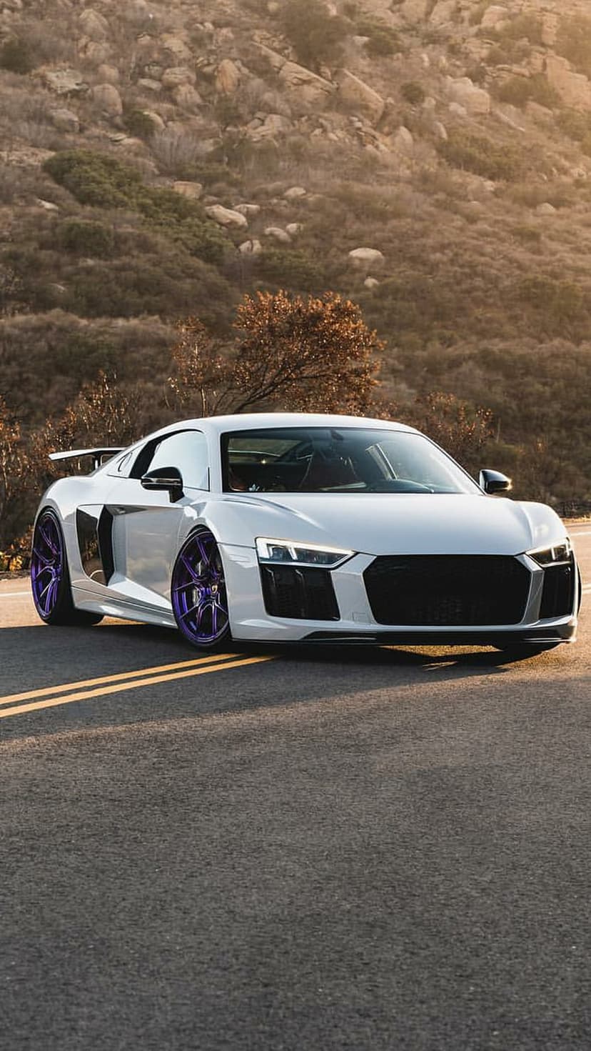 White Audi R8 with purple rims parked on a mountain road during golden hour against a rocky cliffside backdrop.