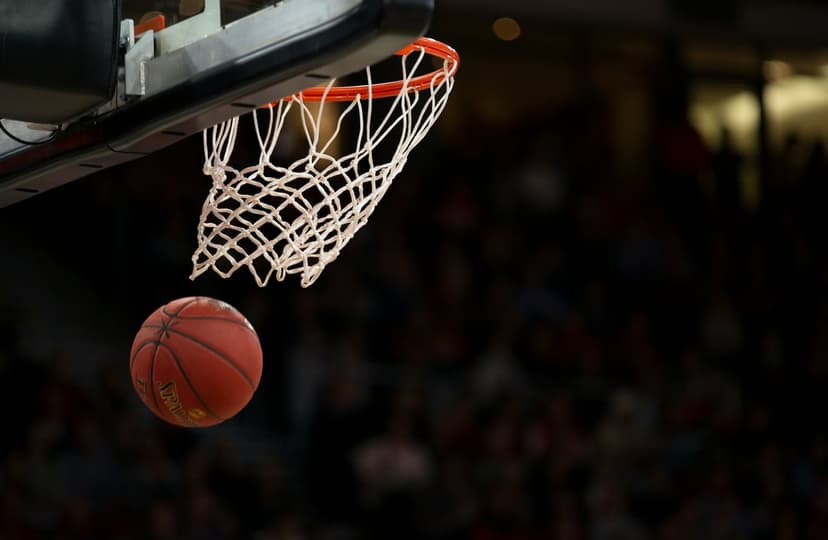 Basketball flying through the air toward an orange rim and white net inside a dark stadium with blurred spectators.