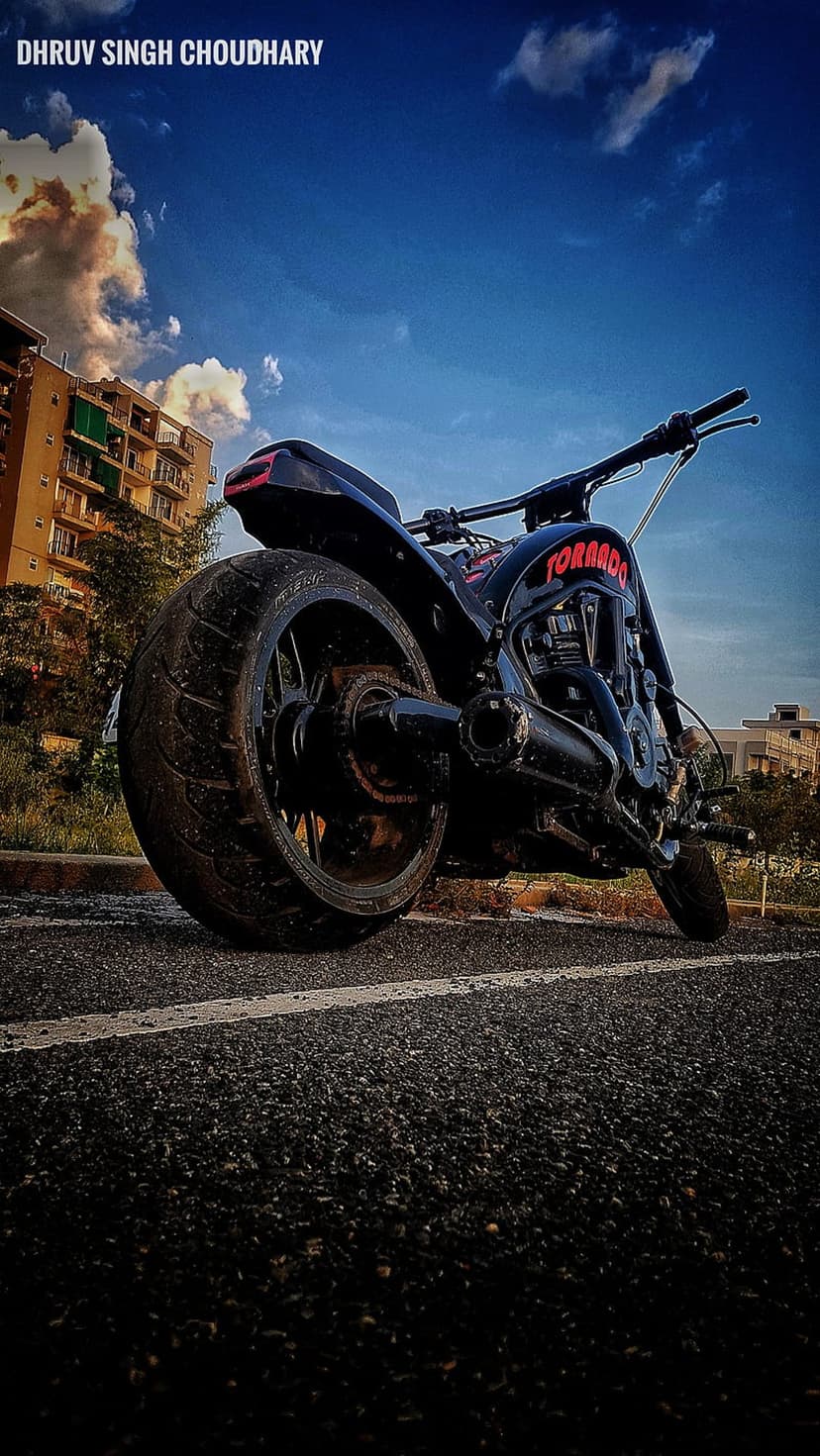 Black Tornado motorcycle with massive rear tire parked on asphalt before a residential building and dramatic sky.