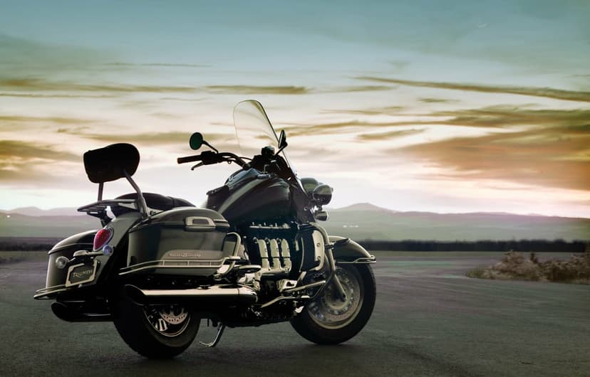 Rear view of a black Triumph Rocket III Touring motorcycle on a road against a soft sunset sky with mountains.