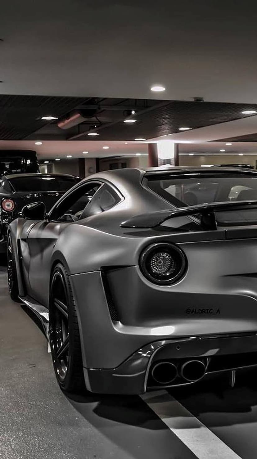 Ferrari F12 rear view in a dark garage featuring matte grey paint, a large spoiler, and round red taillights.
