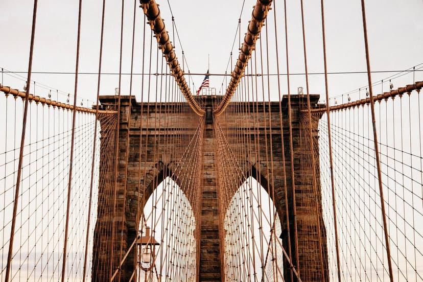 Brooklyn Bridge suspension cables and stone arches with an American flag atop the tower under a muted sky.