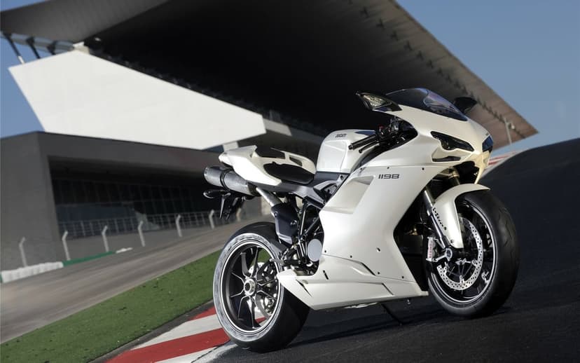 White Ducati 1198 sportbike on a racetrack at a low angle with modern grandstand architecture background.