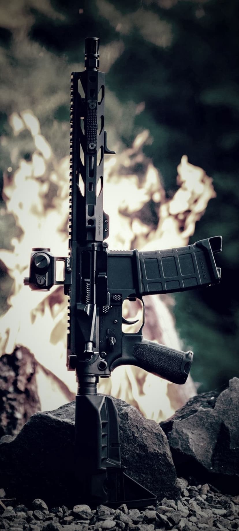 AR-15 rifle with a red dot sight sits on grey rocks with orange flames and green leaves in the background.