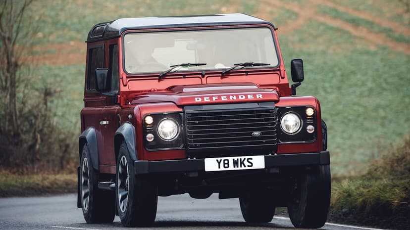 Red Land Rover Defender 90 with glowing headlights driving on a wet asphalt road against a green hillside.