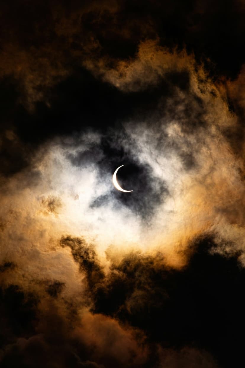 Crescent moon glowing between dark turbulent storm clouds with a fiery orange horizon and deep black sky shadows.