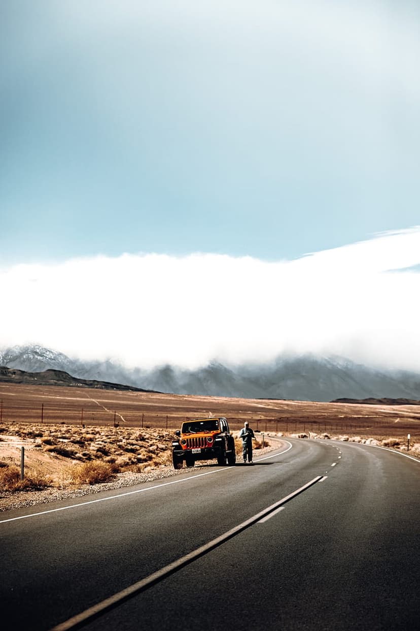 Orange Jeep Wrangler parked on a desert road with a person standing nearby under foggy, distant mountain peaks.