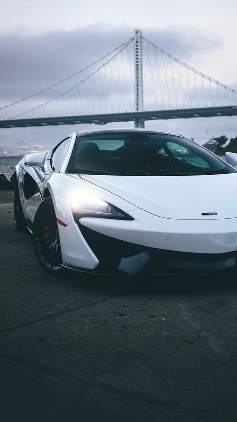 White McLaren sports car with glowing headlights parked on asphalt near a bridge under a cloudy twilight sky.