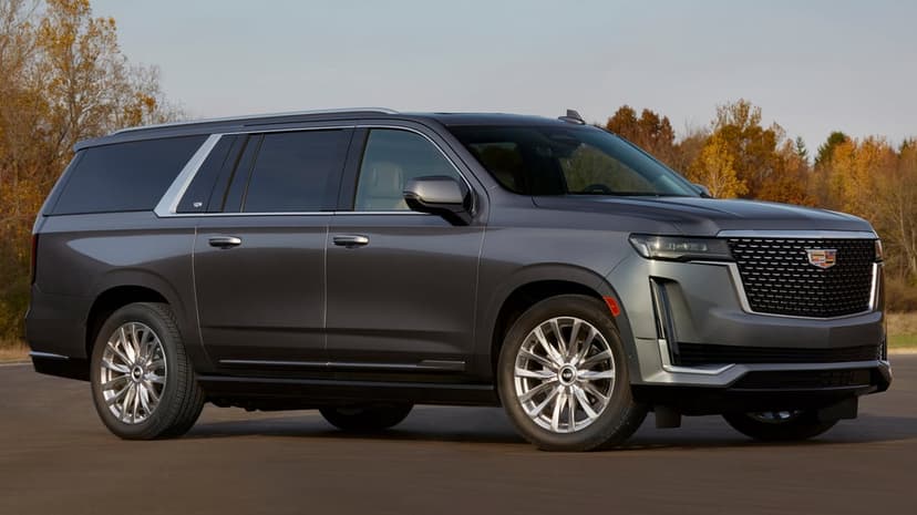 Metallic gray Cadillac Escalade SUV parked on asphalt with vibrant autumn trees and a blue sky in the background.
