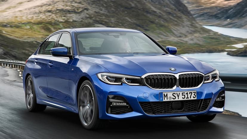 Blue BMW 3 Series sedan driving on a winding mountain road with a scenic lake and rocky hills in the background.