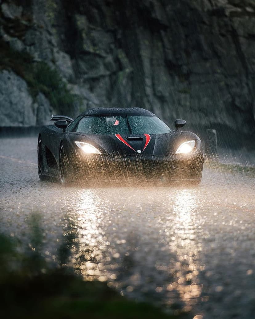Black Koenigsegg hypercar driving on a wet mountain road during heavy rain with bright headlights glowing.