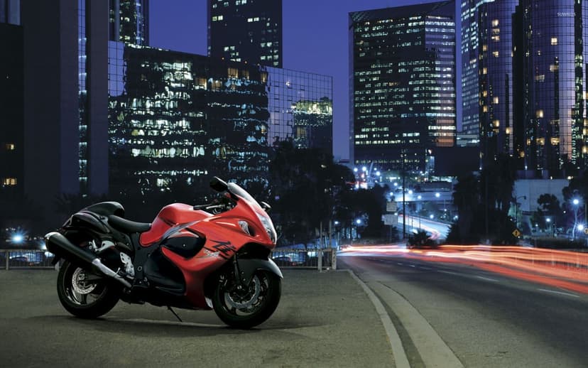 Red and black Suzuki Hayabusa motorcycle parked beside a city highway at night with skyscraper lights and traffic trails.