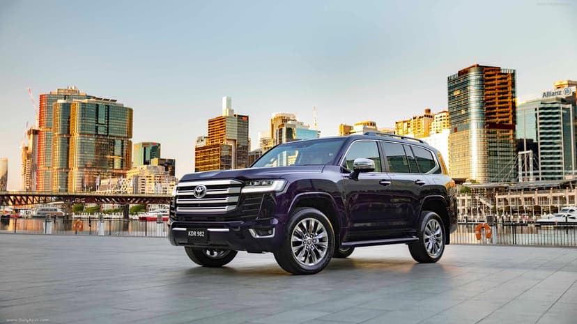 Purple Toyota Land Cruiser 300 parked on a waterfront plaza before a glowing golden hour city skyline.