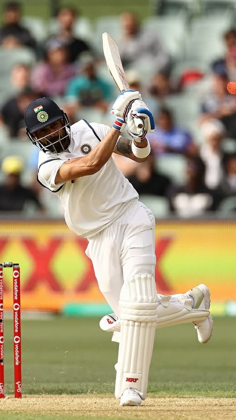 Virat Kohli swings a cricket bat in white test attire on a green pitch with a blurred stadium background.