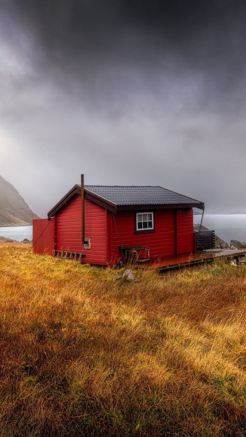 Red wooden cabin stands on golden coastal grass under a heavy overcast sky near the grey northern ocean waves.