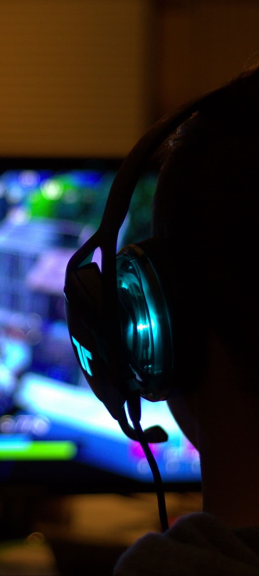 Gamer wearing glowing teal headphones in silhouette against a blurred colorful screen in a dark gaming room.
