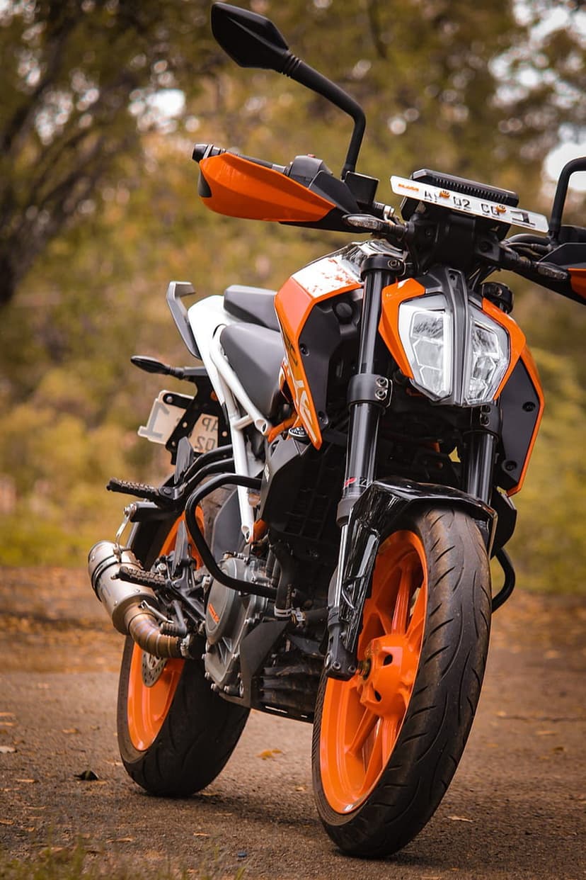 KTM Duke motorcycle with orange wheels parked on a dirt path with blurred green trees in the background.